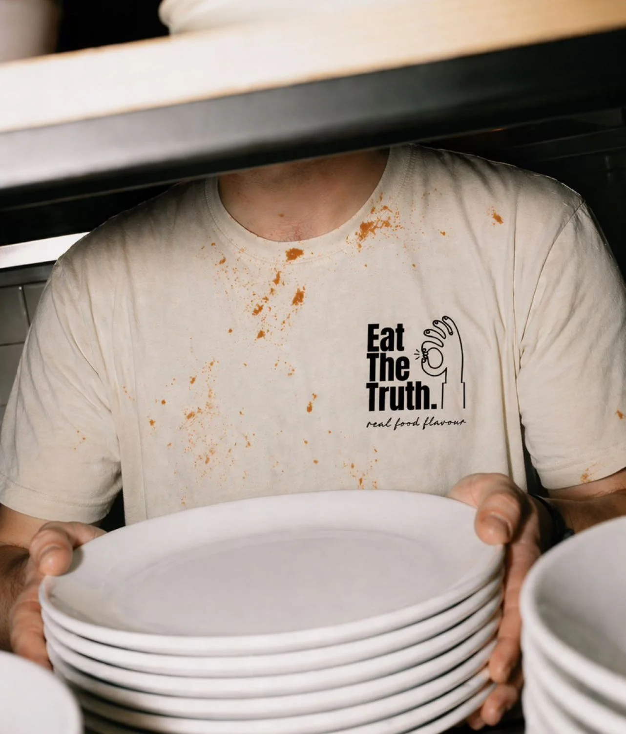 Chef portant un t-shirt Eat The Truth tenant une pile d'assiettes dans une cuisine professionnelle